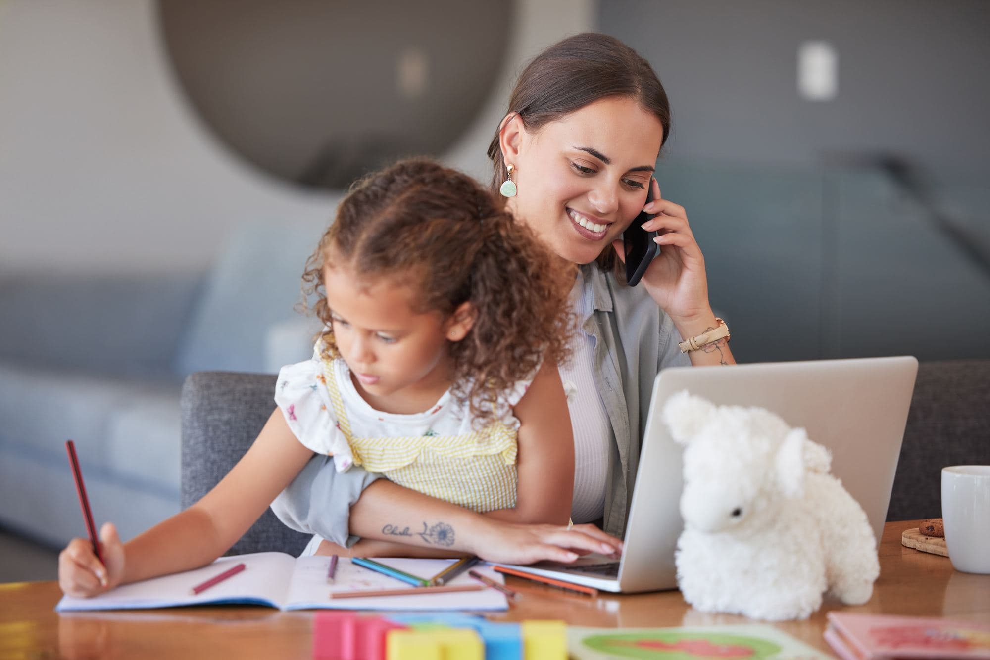 A woman working at a laptop and talking on the phone while her young daughter draws beside her.