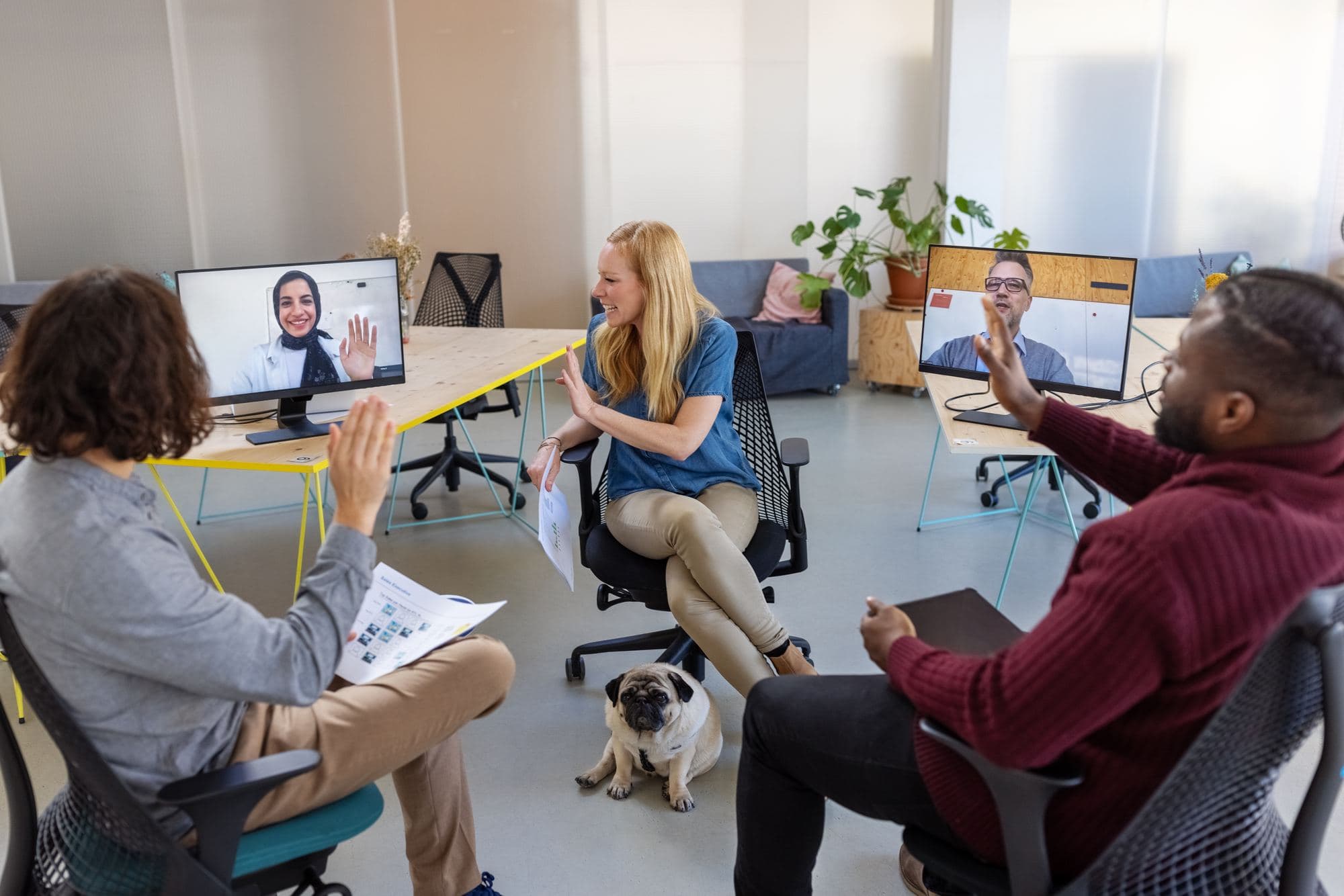 Colleagues in a meeting room talking with two remote participants on screens.